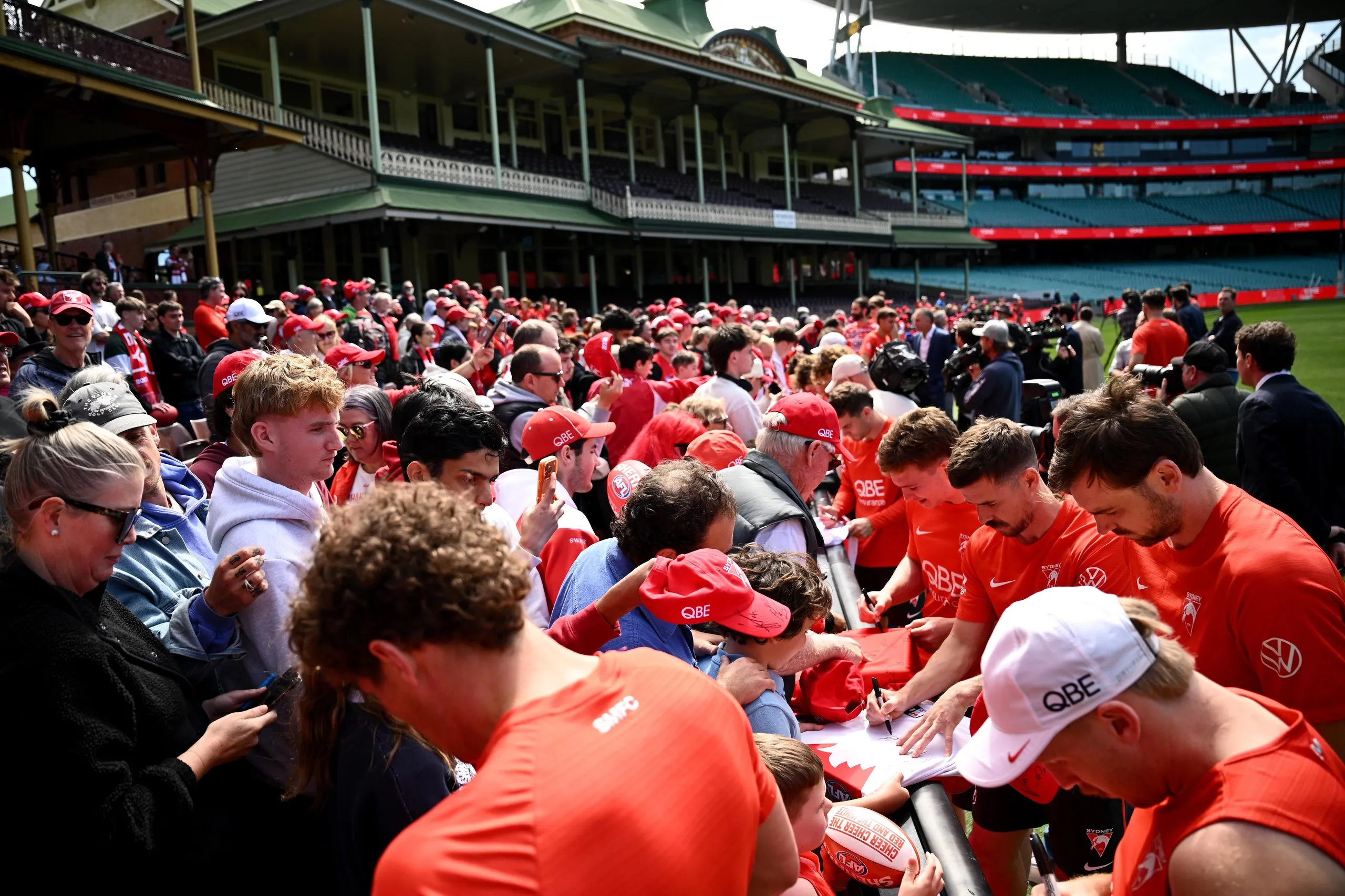 Fans in red and white fill a ground-level stand at the SCG while players standing on the field sign autographs after a training session